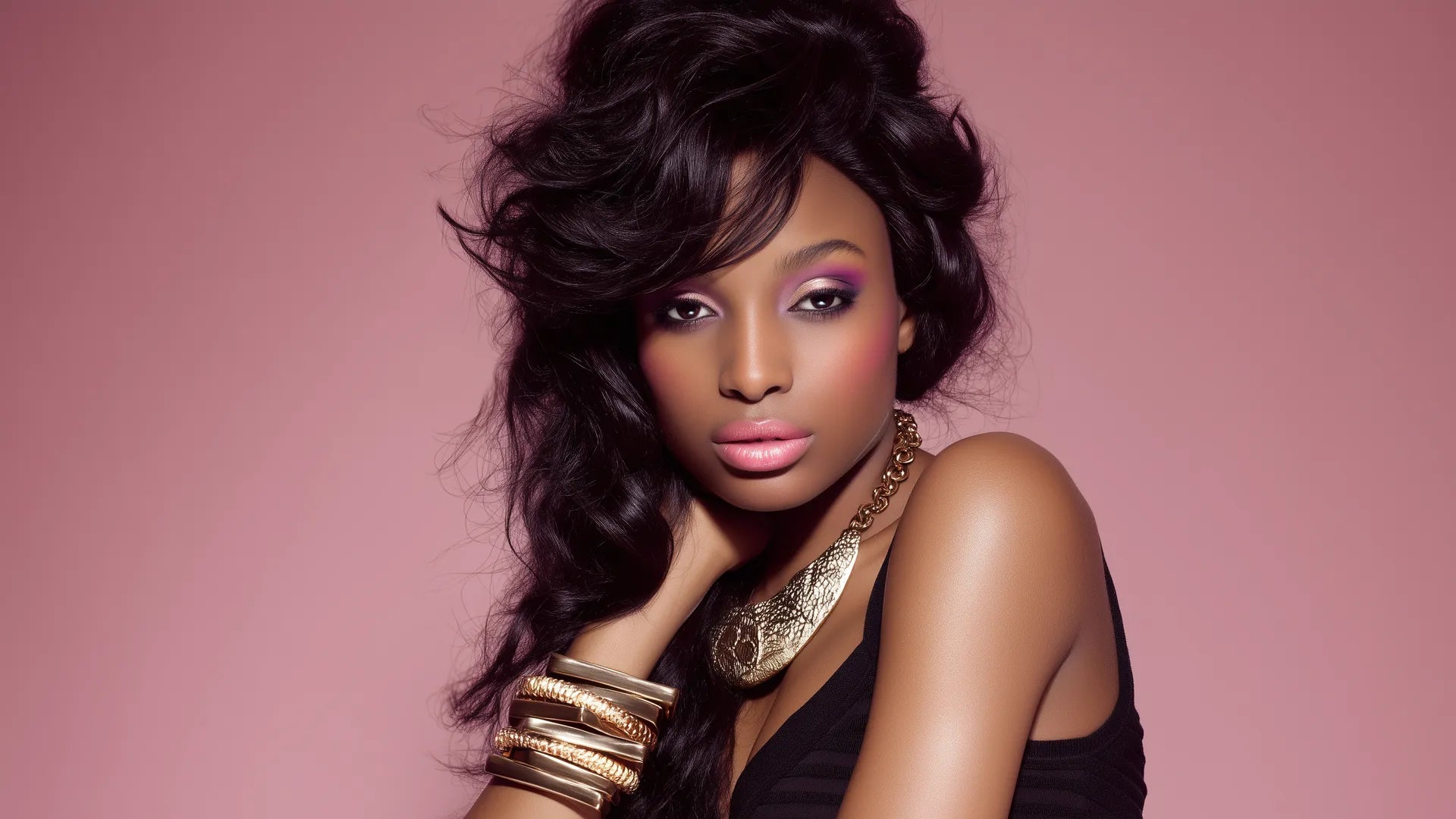 Portrait of a woman with voluminous black wavy hair styled to one side, wearing purple eyeshadow, a black top, and statement gold jewelry including a chunky necklace and stacked bangles, against a dusty rose background