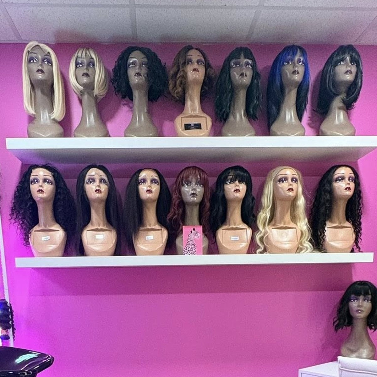 Two showroom shelves lined with wigs in shades from black through brown to blonde, mounted on a salon wash-station wall