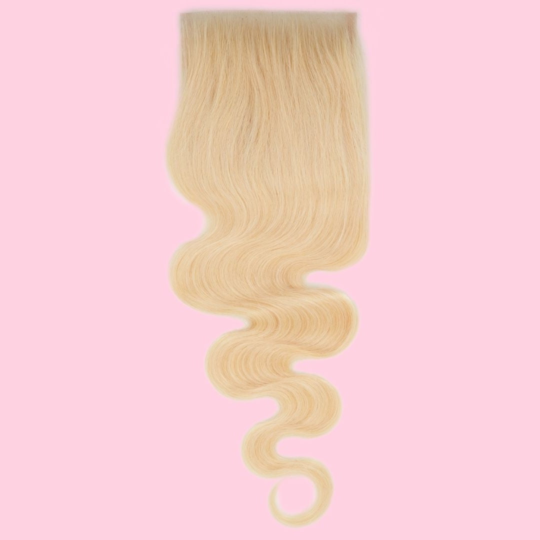 Blonde body wave hair extensions on a transparent lace closure hanging against a pink backdrop with a curl at the tip