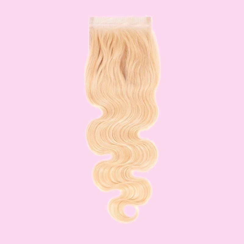 Blonde body wave hair extensions on a transparent lace closure photographed against a pink backdrop with the wave falling in an S curve