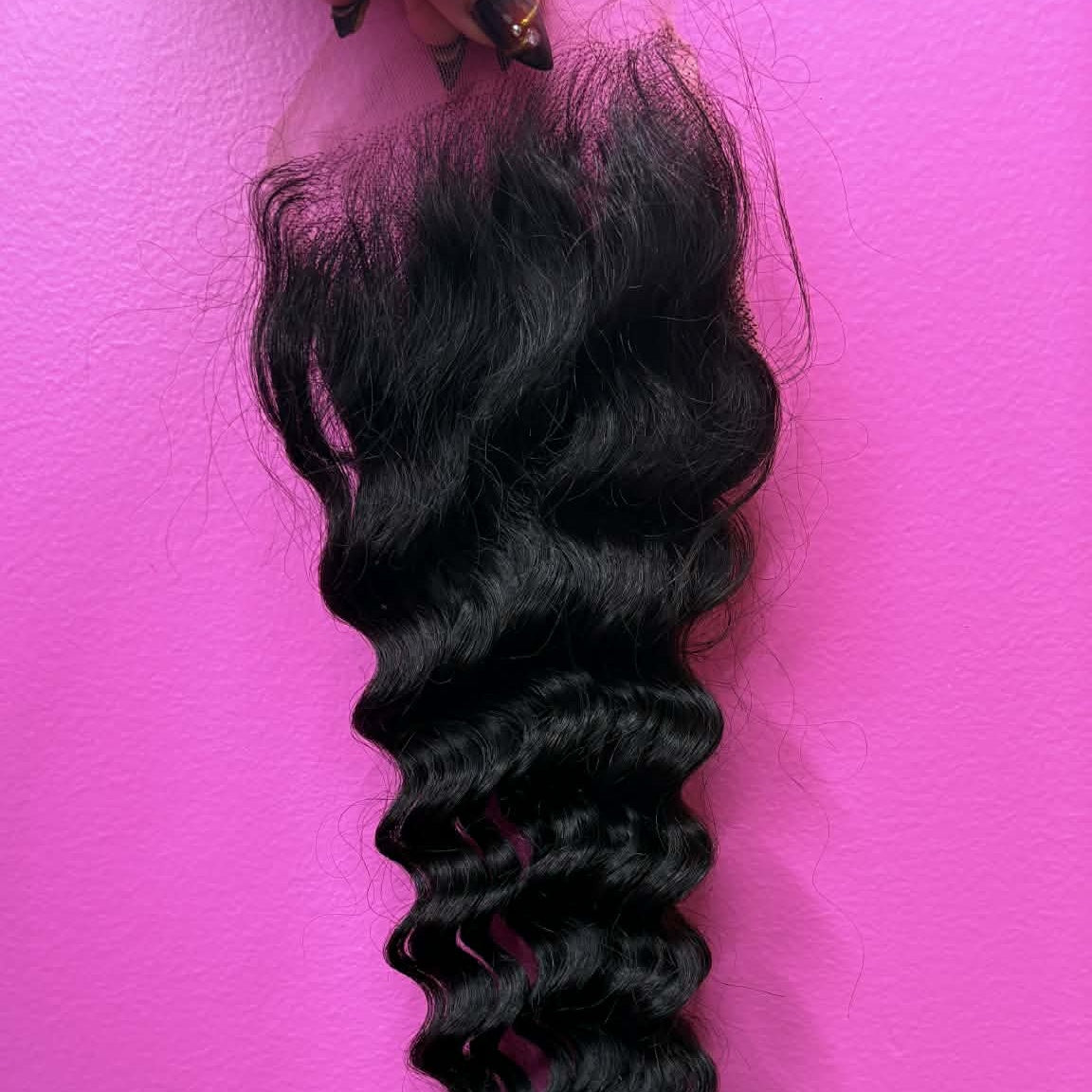 Hand holding 16 inch Brazilian deep wave 4x4 transparent closure hair extension against pink wall showing full curl length from lace to tips