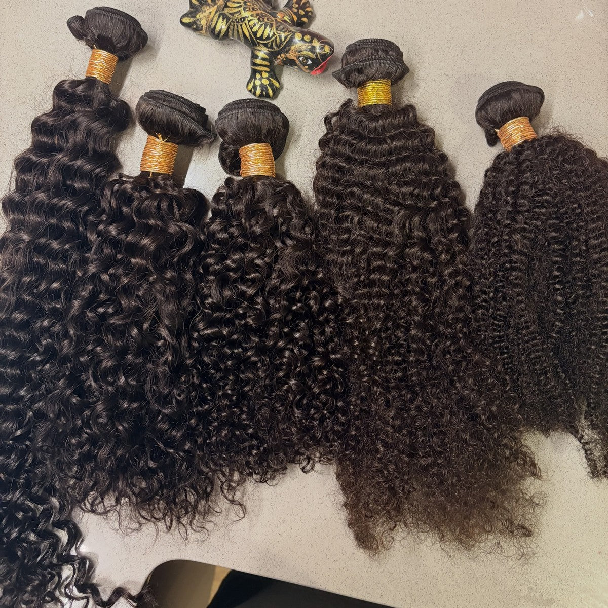 Five natural-black hair bundles laid flat from deep wave to coily, wefts visible at the top, with a small lizard ornament along the upper edge