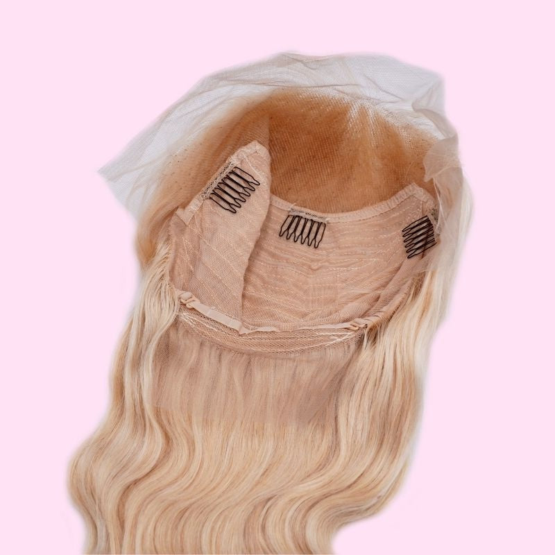 Blonde body wave human hair wig laid flat on pink with the cap opened to show three combs, the mesh dome and the lace front