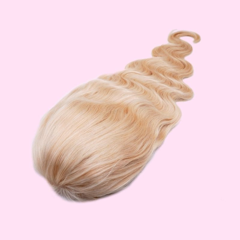 Blonde body wave human hair wig laid crown up on a pink background showing the smooth top and the waved length trailing away