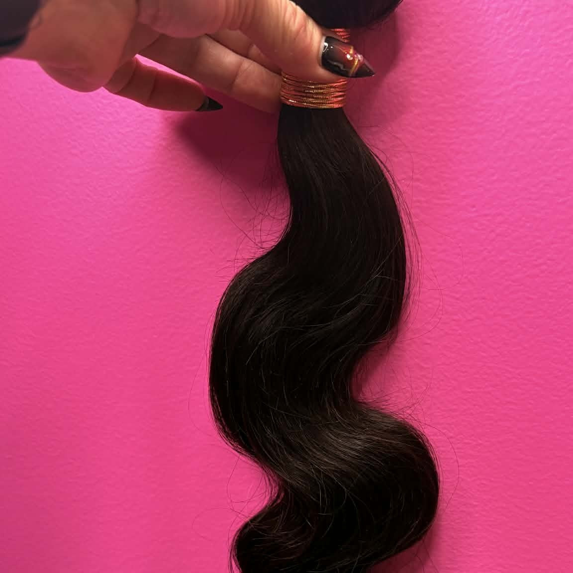 Hand holding 16 inch Brazilian body wave sew-in hair extensions bundle against pink wall showing weft track and flowing S-wave texture