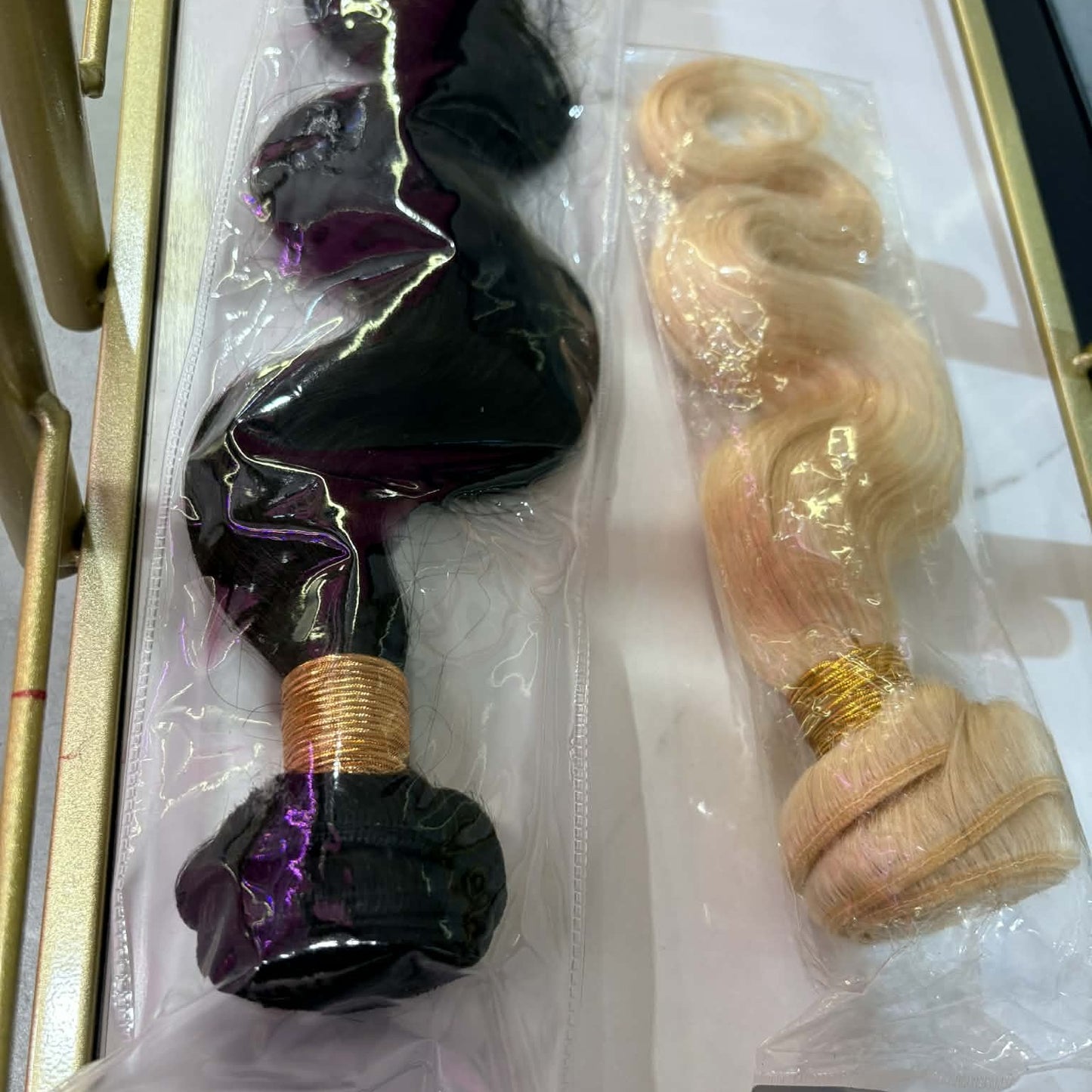 Two body wave sew-in hair extensions bundles in packaging inside gold frame showing natural black and blonde color options