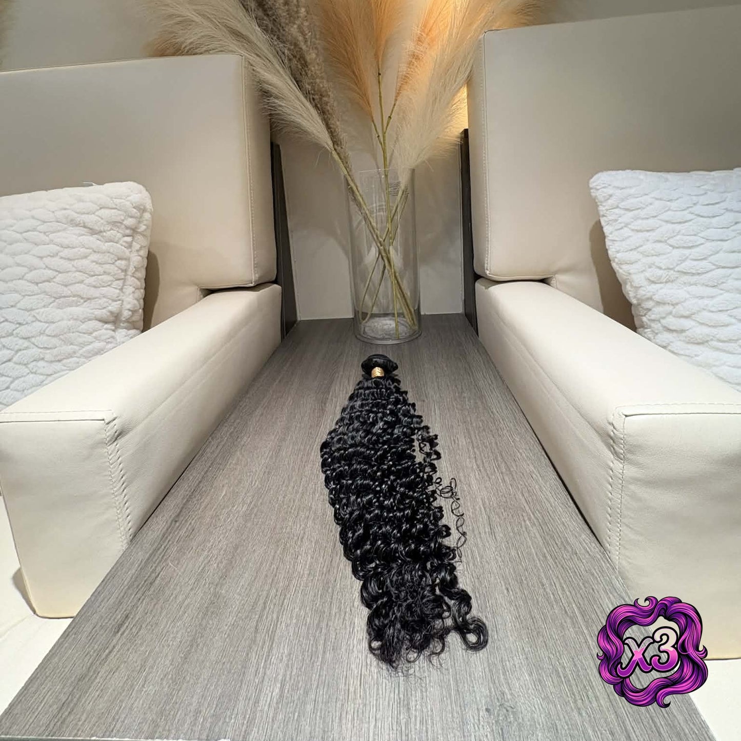 3 bundle deal — 16 inch Brazilian deep wave sew-in hair extensions bundle on table between salon chairs with pampas grass showing full curl length