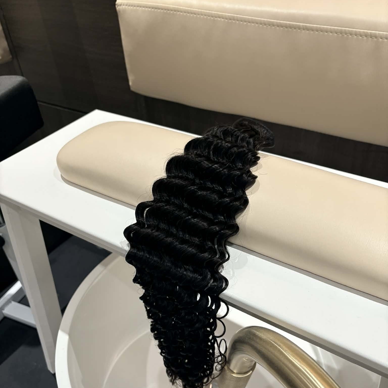 16 inch Brazilian deep wave sew-in hair extensions bundle on salon chair arm showing defined S-spiral curls hanging from weft