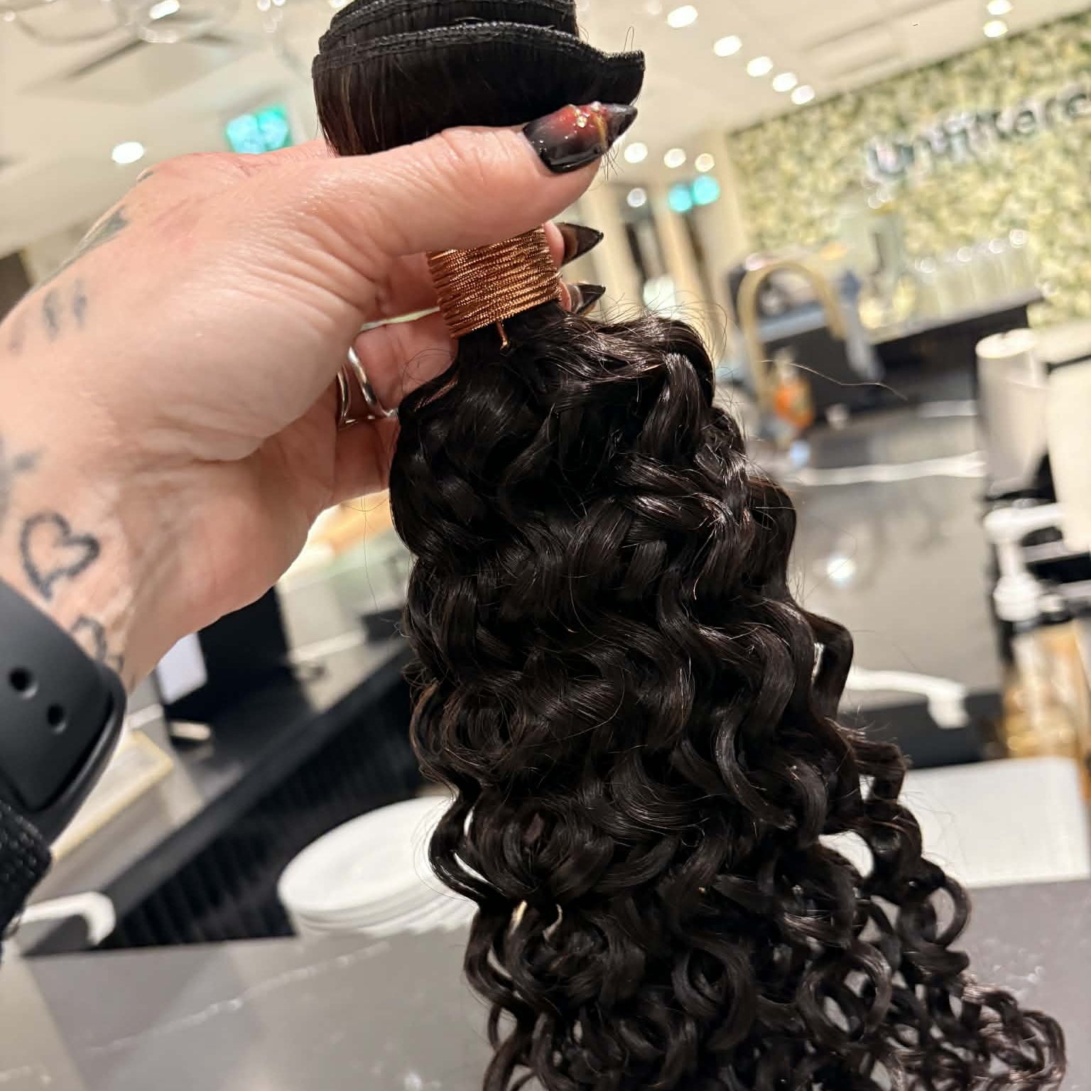 Hand holding 16 inch Brazilian kinky curly sew-in hair extensions bundle in salon showing full curl length and volume from weft to tips