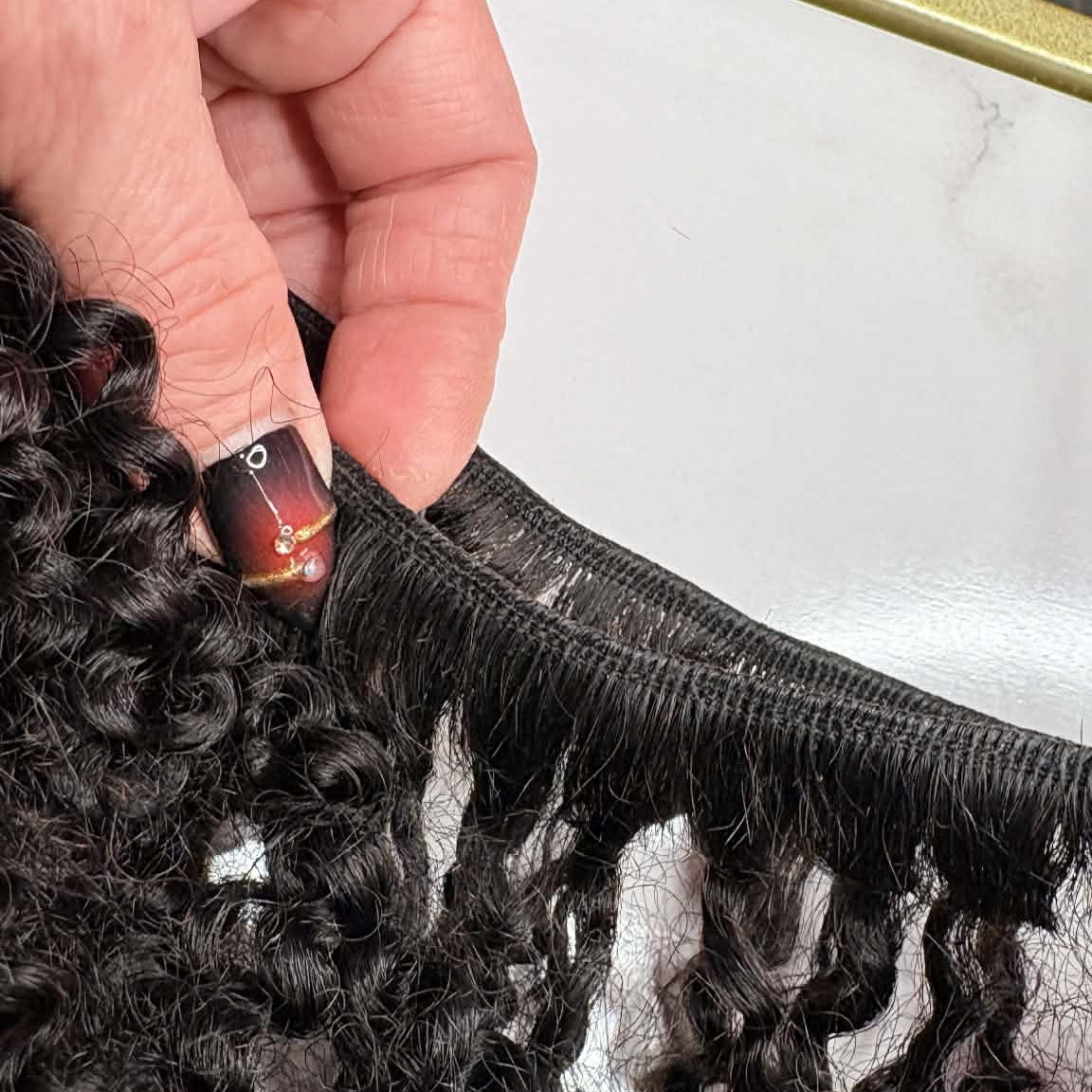 Hand holding 16 inch Brazilian kinky curly sew-in hair extensions bundle showing weft closeup with rolled weft and tight defined curls