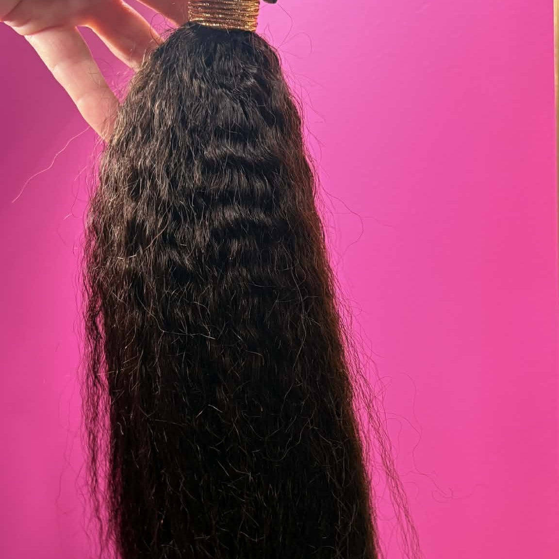 Hand holding 16 inch Brazilian kinky straight sew-in hair extensions bundle against pink wall showing full length and natural volume