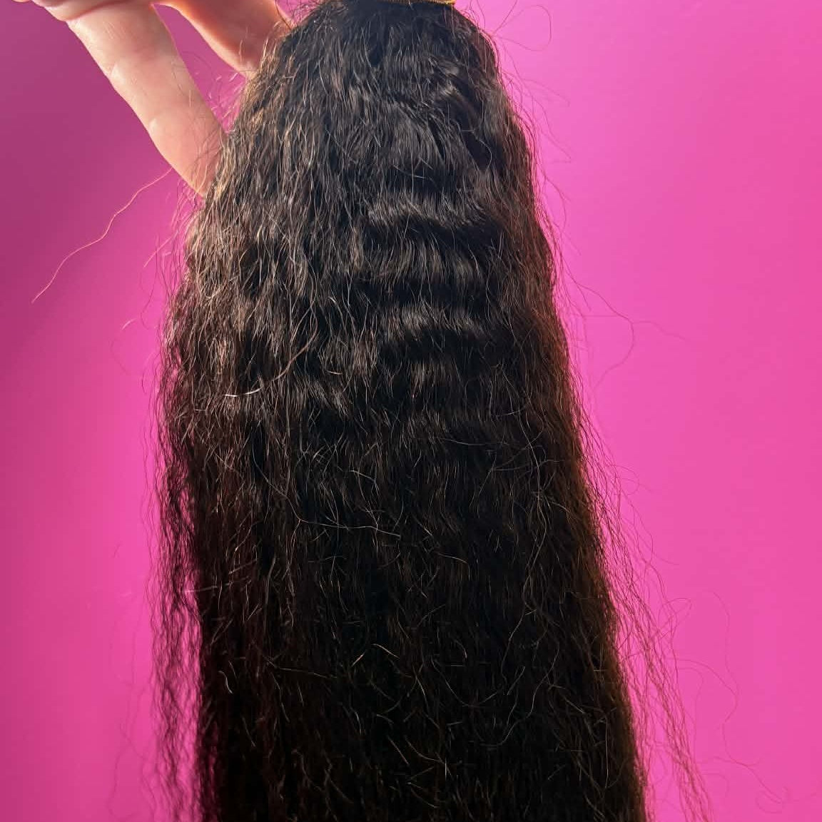 Hand holding 16 inch Brazilian kinky straight sew-in hair extensions bundle against pink wall showing weft and fluffy straight texture