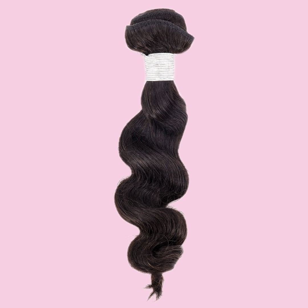 Single bundle of natural black loose wave sew-in hair extensions hanging on a pink backdrop with a white thread wrap around the weft