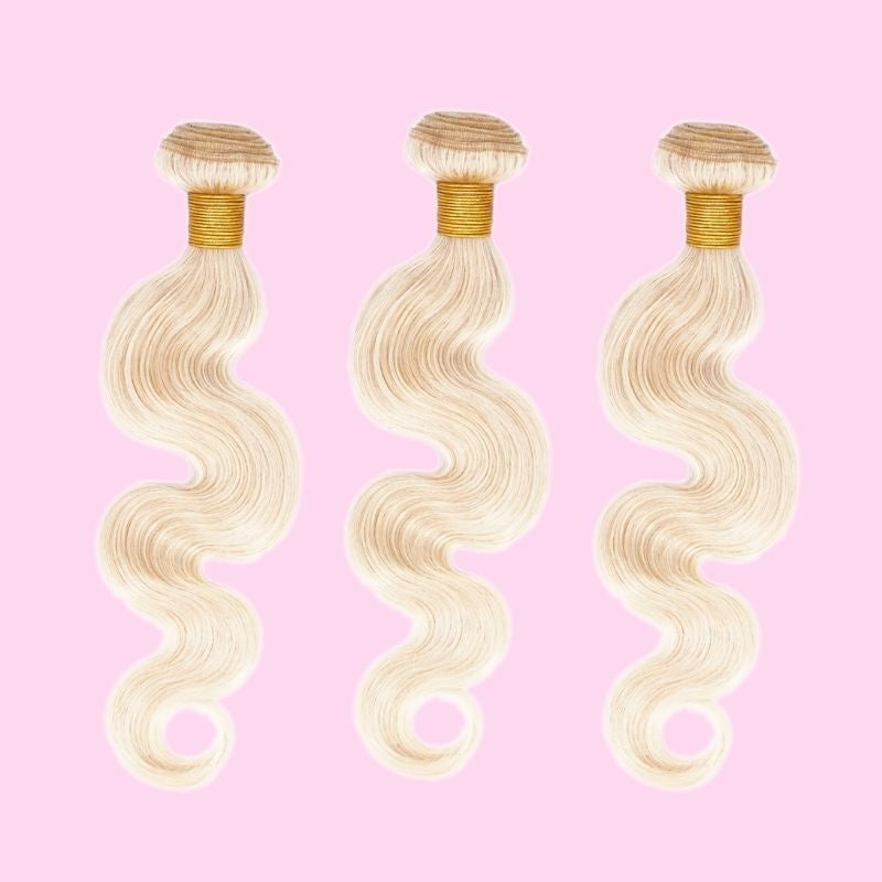 Three bundles of blonde body wave sew-in hair extensions arranged on a pink backdrop with gold thread around each weft